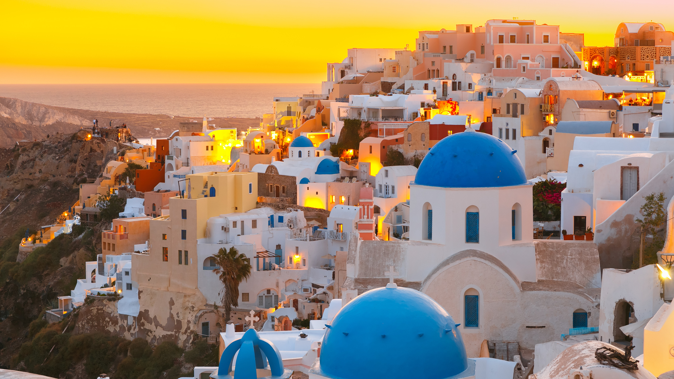 A breathtaking sunset over Santorini, Greece, with golden hues reflecting on the white-washed buildings and the deep blue Aegean Sea."